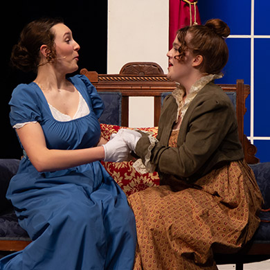 Two actresses on stage looking like they are sharing secrets, smiling at each other