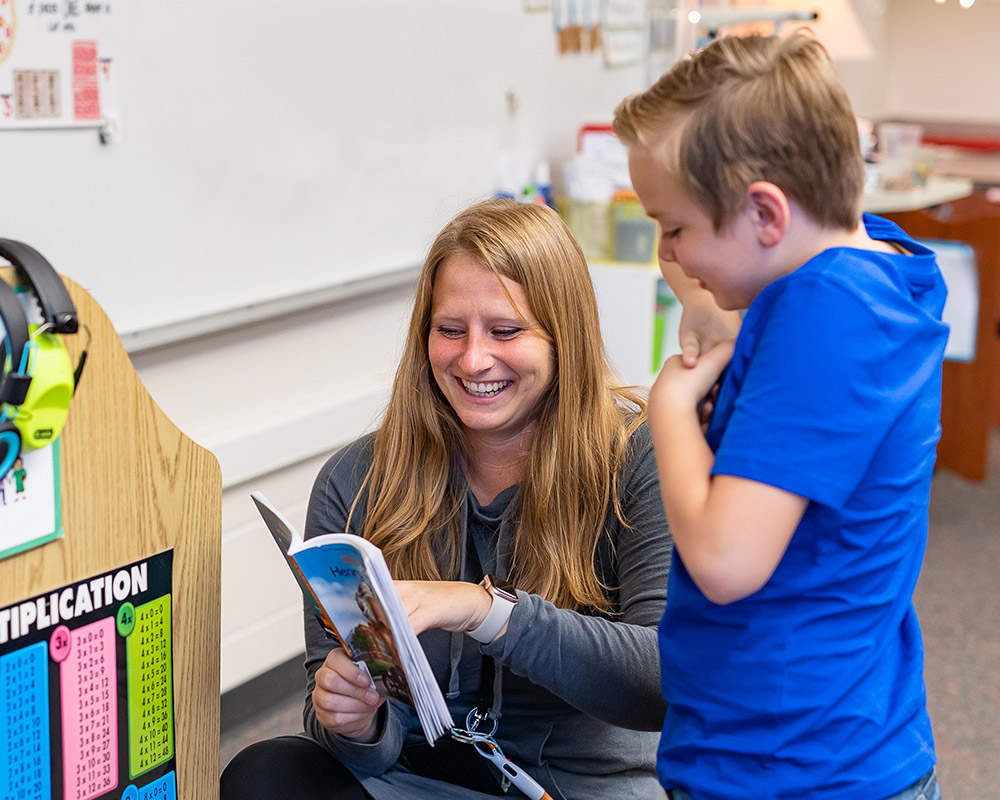 3rd grade boy and teacher smiling together over a book held by the teacher pointing something out