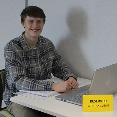 HCHS Spanish Intern Kyle smiling at the camera from his Vita Tax help desk, study materials in front of him.