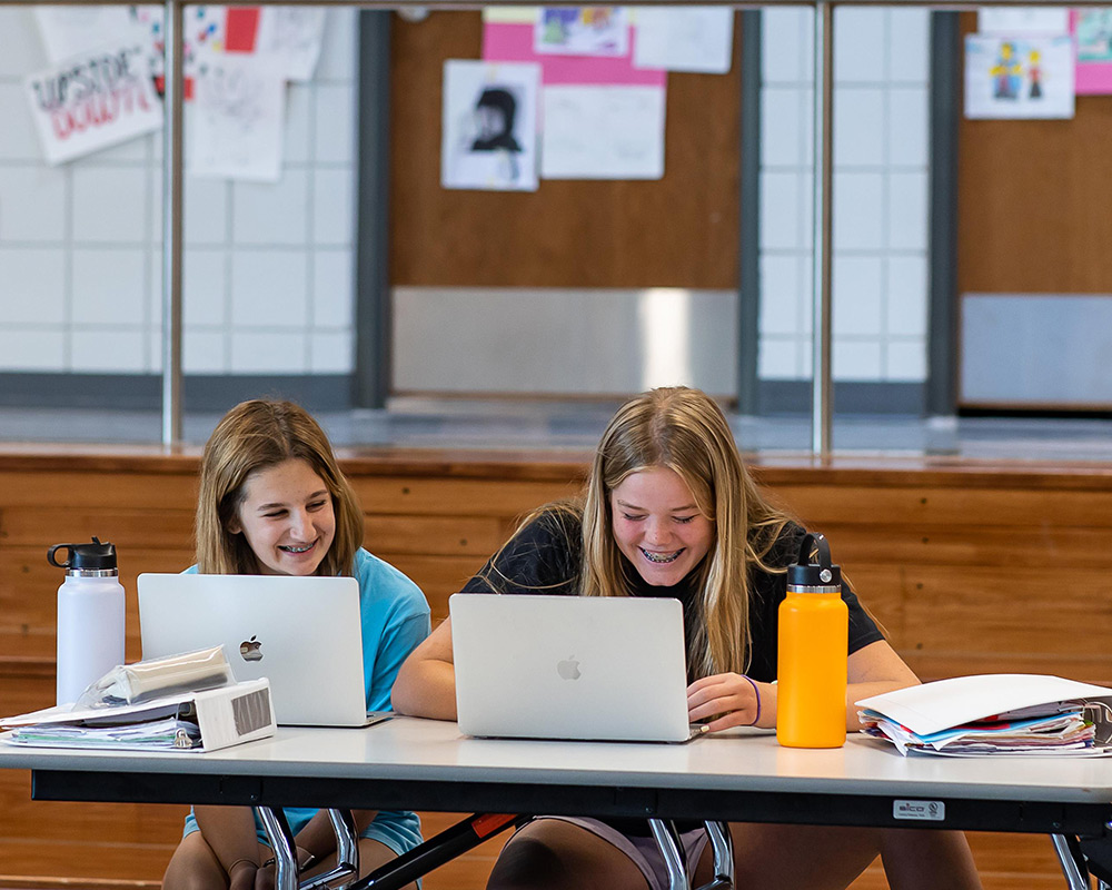2 MS girls smiling over their laptops, working together on school work in the open area.