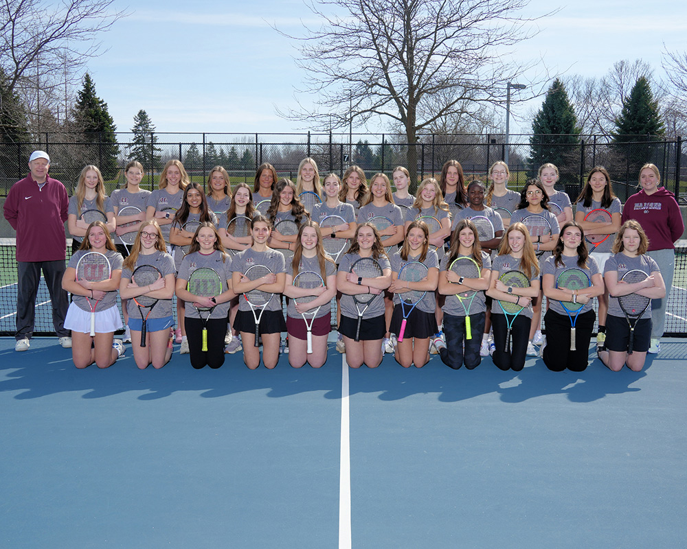 Girls JV tennis team wearing gray uniform tops, while holding their rackets in front of them and smiling at the camera