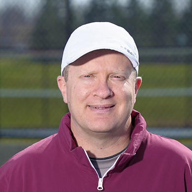 Girls JV tennis coach standing on the tennis courts and smiling at the camera.