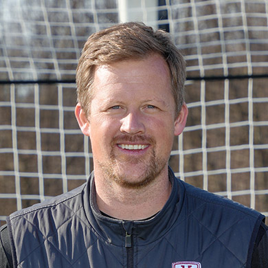 Girls varsity soccer coach standing in front of the soccer goal and smiling at the camera